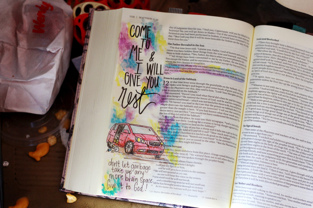 An open bible with a drawing of a van is laying on the floor of a dirty van. There are goldfish crackers and wrappers all around. "Come to me and I will give you rest." is written on the open page.