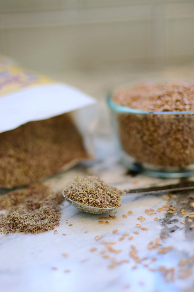 Key ingredients in lactation cookies, ground flaxseed in a measuring spoon with more spilling out of a bag in the background and whole, golden flaxseed scattered in the foreground with a bowl of flaxseed in the background