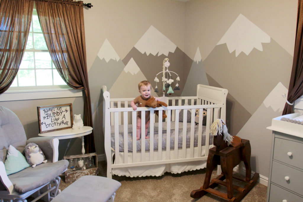 Baby boy standing in crib in a mountain-themed nursery full of diy nursery decor with a rustic nursery sign on the table next to him that reads: There's no buddy like a brother