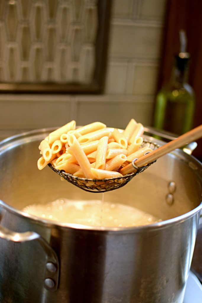 A bamboo skimmer full of one of the ingredients for an easy penne pasta recipe: whole-grain penne cooked in salted boiling water in a stockpot on the stove