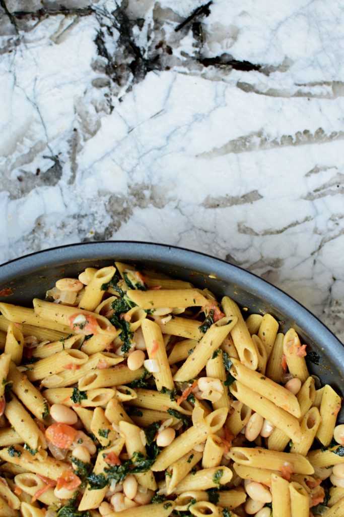A skillet of penne pasta with white beans, kale, tomatoes, onions
