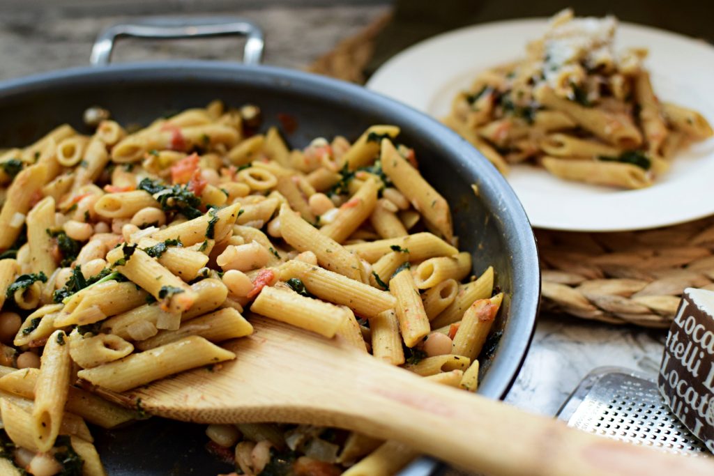 A skillet full of penne pasta with kale, tomatoes, Great Norther beans and a wooden spatula in the pasta