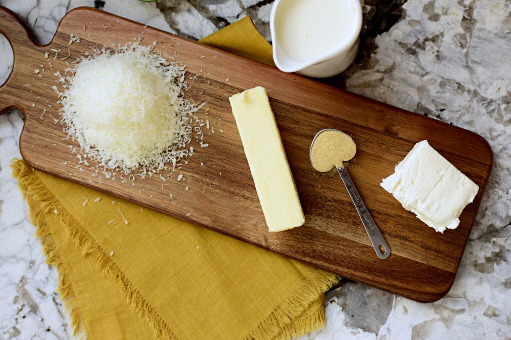 Ingredients for homemade alfredo sauce on a granite countertop, including a mound of freshly grated Parmesan cheese, a stick of butter, granulated garlic in a heart shaped measuring spoon, a block of cream cheese, and a small white cup of heavy cream