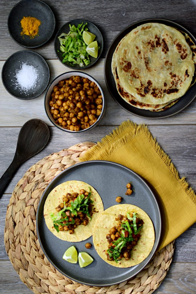 Overhead shot of dinner plate of two vegetarian chickpea tacos on corn tortillas made with Indian chickpeas or garbanzo beans; a bowl of chana masala chickpeas, small plates of salt, turmeric, lettuce and limes, and a plate of flatbread