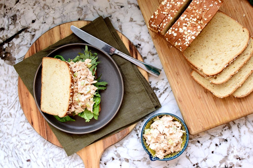 Curry chicken salad sandwich on a plate off to the left, small bowl of curry chicken salad on the bottom of the photo, and a loaf of sliced whole grain bread on the right