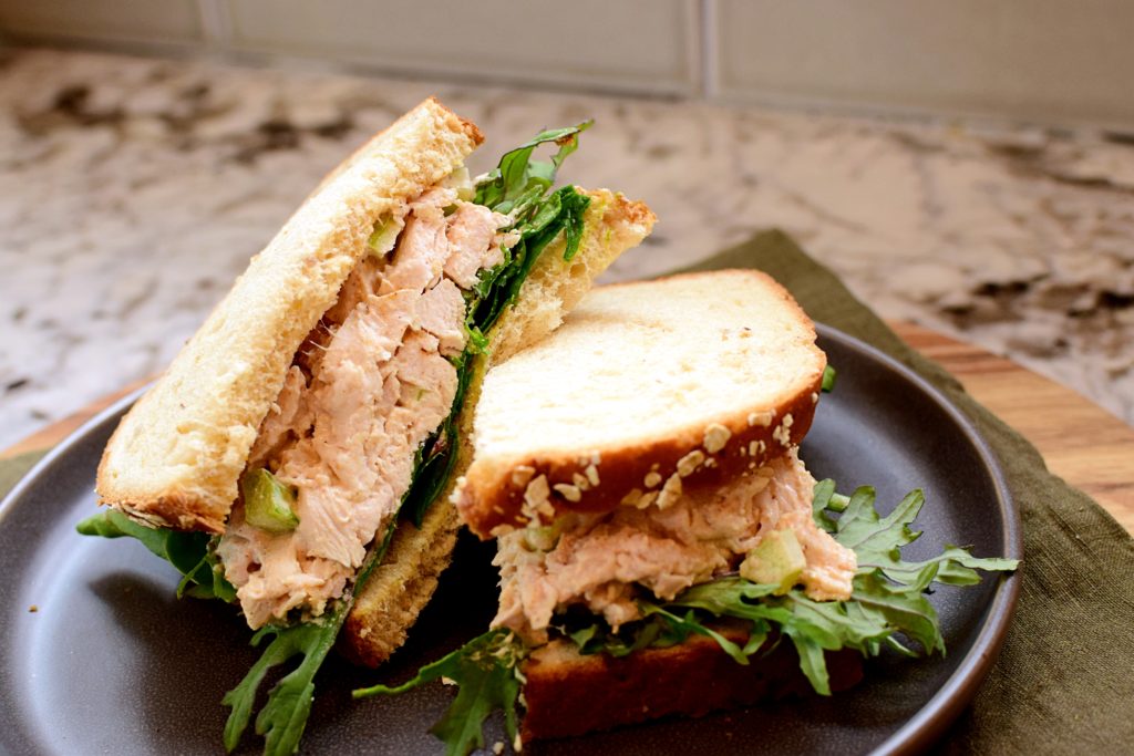 curry chicken salad sandwich cut in half on whole grain bread with a bed of greens on a dark gray plate