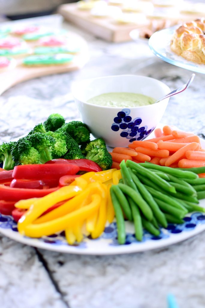 Platter of fresh veggies including broccoli, red pepper, yellow pepper, green beans, carrots and bowl of herby green goddess dip & dressing